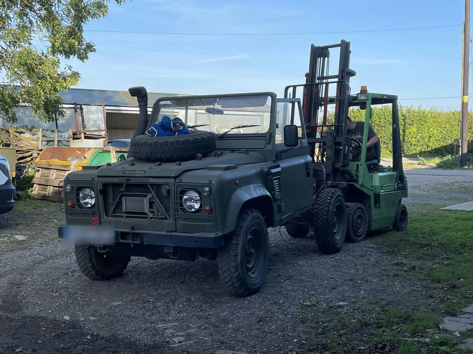 Olive green Land Rover Defender Wolf 90 with a spare wheel on the bonnet, parked beside a forklift